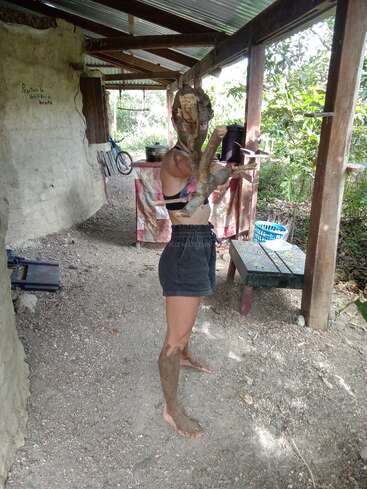 Une personne se tient à l'extérieur sous un abri, pieds nus avec de la boue sur les jambes, tenant des bâtons avec de la boue sur le visage, posant de manière ludique dans un cadre naturel et rustique.