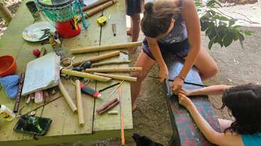 Deux personnes travaillent ensemble en plein air, bricolant avec des bâtons de bambou et des outils sur une table en bois couverte de fournitures, de cahiers et d'objets épars, suggérant un projet de bricolage créatif.