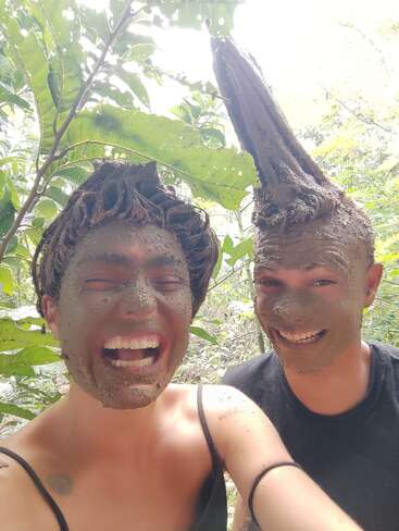 Deux personnes se couvrent joyeusement le visage et les cheveux de boue, créant ainsi des coiffures ludiques et créatives. Elles rient ensemble en plein air, entourées de feuilles vertes et de la lumière du soleil dans la nature.
