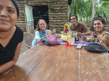 Quatre personnes sont assises à une table rustique en bois à l'extérieur ; elles sourient et partagent un repas. Le cadre est chaleureux, avec des arbres et une cabane en rondins en arrière-plan.
