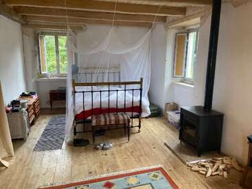 La imagen muestra un acogedor dormitorio con cama, estufa de leña y suelo de madera, con cama con dosel, cómoda y alfombra, con una serena vista de los árboles al otro lado de la ventana.