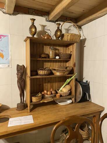 Un estante de cocina de madera sostiene cestas, cebollas, una calabaza, jarrones decorativos y varios recipientes. Cerca, sobre una mesa rústica, hay una estatua de madera tallada y utensilios de cocina.