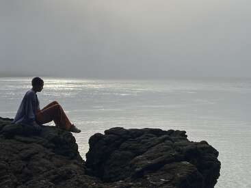 Una persona está sentada tranquilamente sobre unas rocas oscuras, contemplando una masa de agua tranquila y brumosa bajo un cielo gris. La escena es apacible y contemplativa.