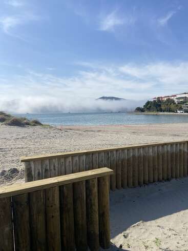 Una playa de arena con una barrera de madera en primer plano, aguas azules tranquilas, niebla sobre colinas lejanas y casas enclavadas en una ladera verde. Cielo despejado.