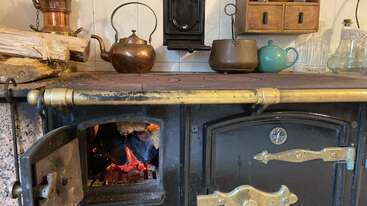 Esta imagen muestra una antigua estufa de leña con fuego en su interior. Encima hay varias teteras, jarras y utensilios de cocina que crean un ambiente acogedor.