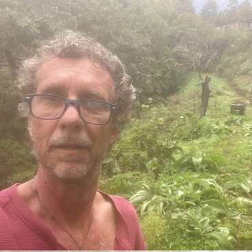 Un hombre con gafas y una camiseta roja se hace un selfi en un jardín frondoso y descuidado. Al fondo, otra persona trabaja cerca de una carretilla.