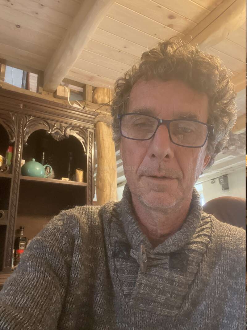 La imagen muestra a un hombre de pelo rizado y gafas, con un jersey gris, delante de un armario de madera con estanterías y un techo blanco con vigas vistas.