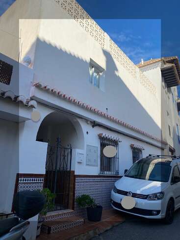 This image shows a white, two-story Mediterranean-style house with patterned tiles, black iron gate, and potted plants. A white Volkswagen van is parked outside.
