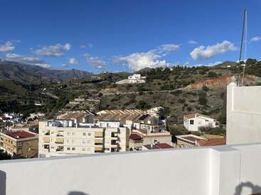 This image shows a sunny hillside town with white buildings, terracotta roofs, and green hills in the background under a bright blue sky with clouds.