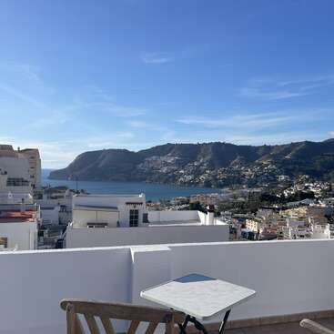 A rooftop terrace with a wooden chair and table overlooks a coastal town, the sea, and scenic hills under a clear blue sky with light clouds.