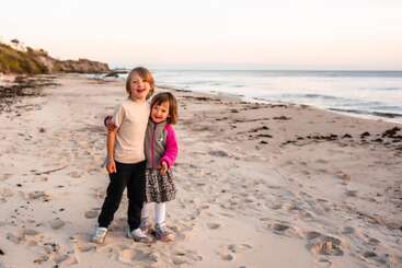 Dos niños pequeños están de pie en una playa de arena, sonriendo y abrazándose. El sol se está poniendo, creando un ambiente tranquilo y cálido junto al océano.