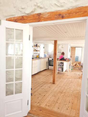 Una acogedora cocina rústica con suelos y vigas de madera muestra a una persona trabajando en una isla. La luz natural que entra por las puertas de marco blanco crea un ambiente cálido.