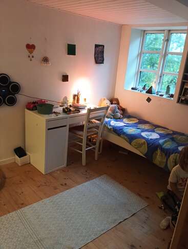 Un dormitorio acogedor y ordenado con un escritorio con una silla, arte en la pared, cama con ropa de cama azul, ventana grande, animales de peluche, alfombra y suelo de madera. Niño sentado.