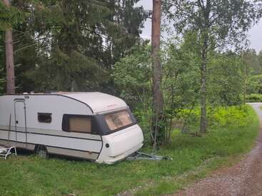 Ein weißer Wohnwagen steht auf einer Wiese neben einer Schotterstraße, umgeben von hohen grünen Bäumen und üppigem Wald, was eine friedliche, abgeschiedene Atmosphäre schafft.