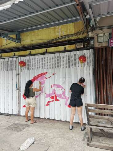 Dos mujeres están pintando un mural de un pez koi en una verja metálica blanca. Por encima cuelgan farolillos chinos. La escena parece estar al aire libre.