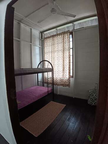 Una habitación pequeña y sencilla con suelo de madera, paredes blancas, un ventilador de techo, una litera con colchón rosa, una ventana con cortinas de flores, una cesta y una alfombra estampada.