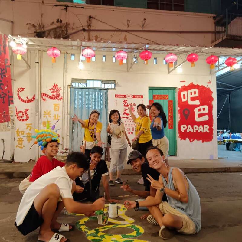 Un grupo de gente alegre está pintando un colorido mural de una serpiente en la calle, rodeado de farolillos y llamativas decoraciones chinas, mientras celebran juntos por la noche.