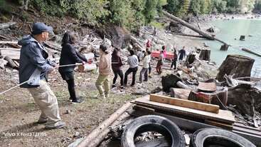 Un grupo de personas trabaja en equipo, tirando de una cuerda en la orilla rocosa de un lago rodeada de madera flotante, árboles y provisiones, lo que pone de manifiesto el espíritu de equipo y el esfuerzo colectivo.