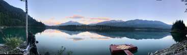 Un tranquilo lago de montaña refleja el cielo y los picos lejanos. Los árboles enmarcan la escena, con un pequeño embarcadero rojo flotando sobre las aguas cristalinas y tranquilas.