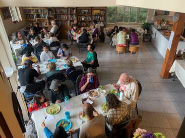 Un grupo variado de personas se reúne en una acogedora sala repleta de libros, sentadas alrededor de las mesas, compartiendo una comida, charlando y disfrutando de la compañía mutua en un ambiente cálido.