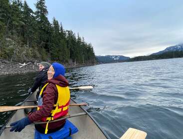 Dos personas reman en una canoa por un lago tranquilo, rodeadas de colinas boscosas y montañas lejanas. Visten ropa de abrigo y llevan chalecos salvavidas, y sonríen felices.
