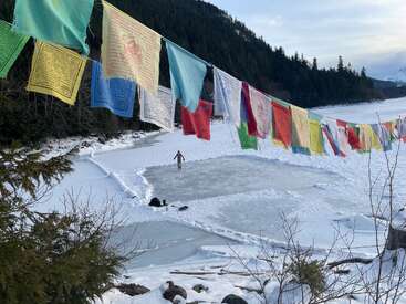 Banderas de oración de colores cuelgan sobre un lago helado, rodeado de nieve, bosques y montañas. Una persona se encuentra de pie en una pista de hielo despejada, disfrutando de la belleza del invierno.