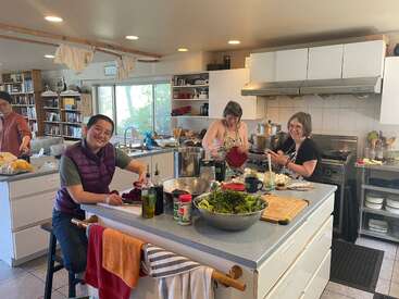 Cuatro personas preparan alegremente la comida juntas en una cocina espaciosa y bien iluminada. Se pueden ver ingredientes frescos, utensilios de cocina y paños, lo que crea un ambiente cálido y acogedor.