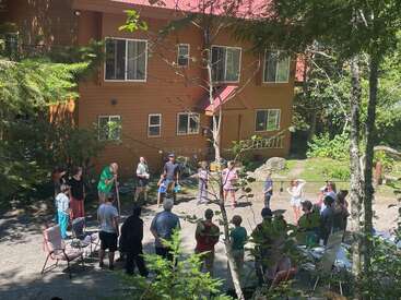 Un grupo de personas, entre las que hay niños y adultos, se ha reunido frente a una casa de madera rodeada de árboles, disfrutando de un día soleado, quizá durante un evento social.