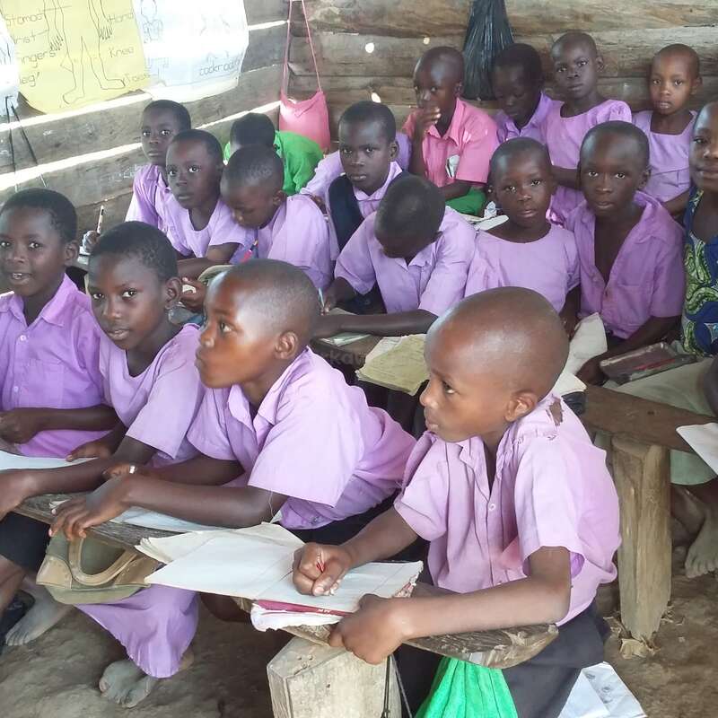 The image shows a group of children sitting at desks in a classroom, with a few adults standing in the background, likely teachers or staff members. The children are engaged in an activity.