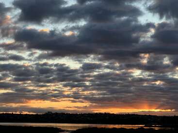 Dark, dramatic clouds blanket the sky at sunset, with golden and orange tones glowing near the horizon, reflecting softly on a calm lake below. Peaceful scenery.