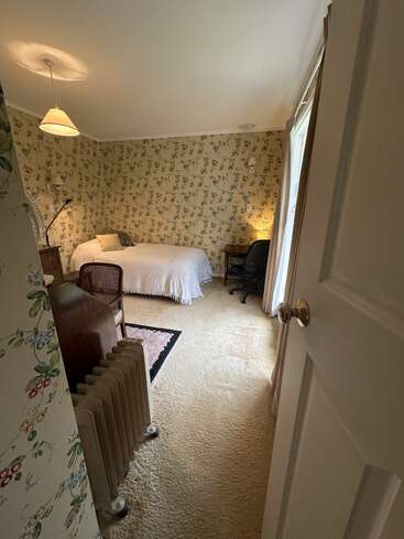 A cozy bedroom with floral wallpaper, a single bed, desk, chair, lamp, and radiator. Soft carpeting covers the floor, and sunlight enters through curtains.