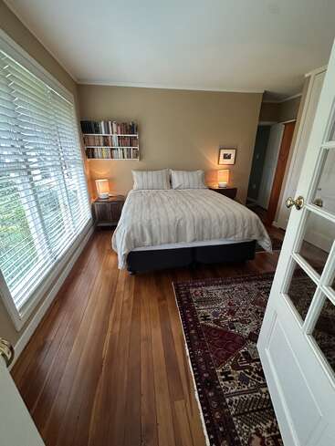 This cozy bedroom features a neatly made bed, wooden floors, a large window with blinds, bedside lamps, bookshelves, framed art, and an ornate area rug.