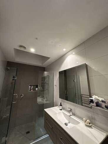 This modern bathroom features a glass shower, large mirror, sleek vanity with white countertop, overhead lighting, and wall tiles. A soap dispenser and tissue box are visible.