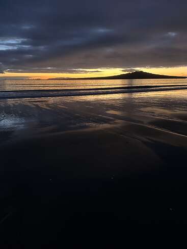 A dramatic sunset over a tranquil beach, with dark clouds looming above, golden sunlight reflecting on wet sand, and a distant island on the horizon. Peaceful atmosphere.