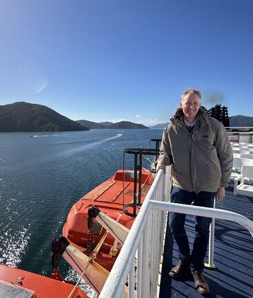 A man stands smiling on a ferry deck, overlooking calm blue water and green hills. It’s sunny, with clear skies and a bright orange lifeboat nearby.