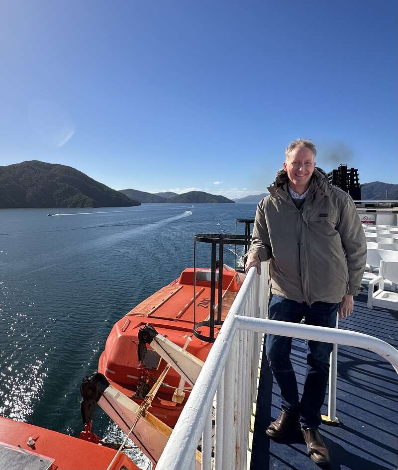 A man stands smiling on a ferry deck, overlooking calm blue water and green hills. It’s sunny, with clear skies and a bright orange lifeboat nearby.