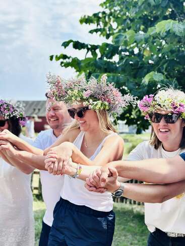 Eine Gruppe von Menschen mit Blumenkränzen und Sonnenbrillen, die draußen fröhlich Händchen halten und lächeln. Es sieht aus wie ein festliches Sommerfest oder ein Treffen unter strahlend blauem Himmel.
