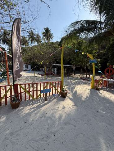 A sunny tropical beach scene with white sand, palm trees, colorful flags, potted plants, a wooden fence, and rustic structures in a relaxed, inviting atmosphere.