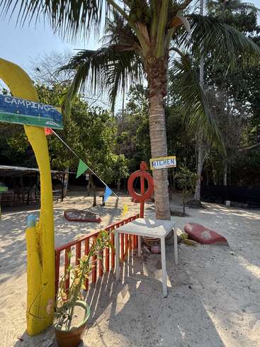 A sandy outdoor campsite with palm trees, colorful signs reading "CAMPING" and "KITCHEN," potted plants, a table, festive flags, and relaxed tropical atmosphere.