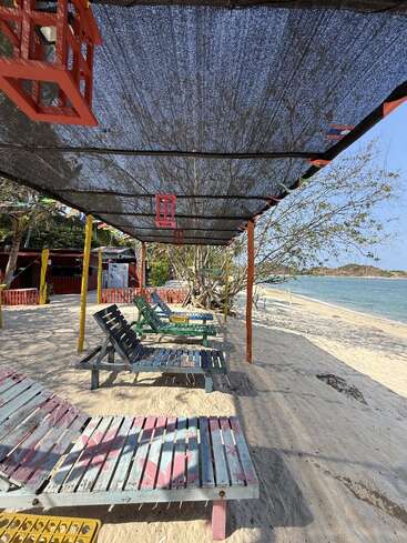 Colorful wooden lounge chairs sit under a shaded canopy on a sandy beach. Red lanterns hang above. The sea and trees create a peaceful, tropical atmosphere.