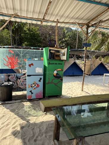 A beachside setup features painted cabinets labeled "Books and Games," a glass-top table, sand, colorful tents, palm trees, and a shaded roof with wooden supports.