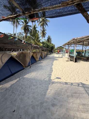 Several tents are set up on a sandy beach under a shaded canopy, surrounded by palm trees, colorful flags, and rustic structures, creating a tropical camping atmosphere.