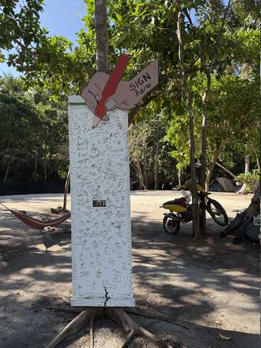 A tall white signing board stands outdoors, covered with signatures. Above it, a cartoon hand holding a pencil points to the words “SIGN here.” Hammock and motorcycle nearby.