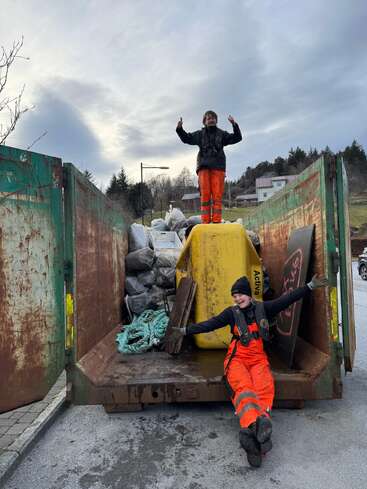 Two people in orange work overalls pose happily on a large container filled with collected trash, ropes, and debris, possibly after a successful cleanup effort outdoors.