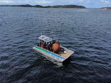 A small boat with five people, possibly divers, floats on open water. There is a large rusty object on board. Islands are visible in the background.