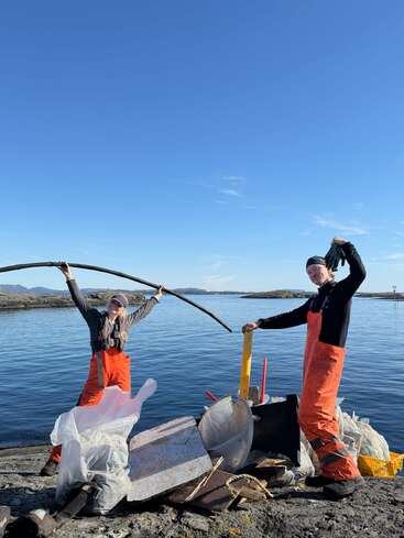 Two people in orange overalls stand by a pile of collected trash near the water, celebrating their cleanup effort under a clear blue sky and sunshine.