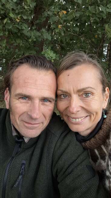 A smiling couple poses closely together for a selfie outdoors, surrounded by green trees. They both wear warm clothing and appear happy, enjoying a moment in nature.