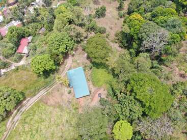 This aerial image shows a single blue-roofed house surrounded by lush green trees, fields, and vegetation, with a dirt road and nearby scattered rural buildings.
