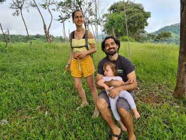 A woman stands smiling in a grassy field while a man sits barefoot, holding a young, sleepy child. Trees and hills create a beautiful, lush backdrop.