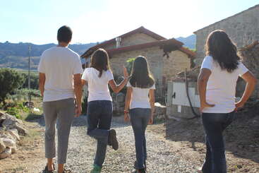 Four people, dressed in white shirts and jeans, walk towards a rustic stone house in a rural setting, with mountains and greenery visible in the background.